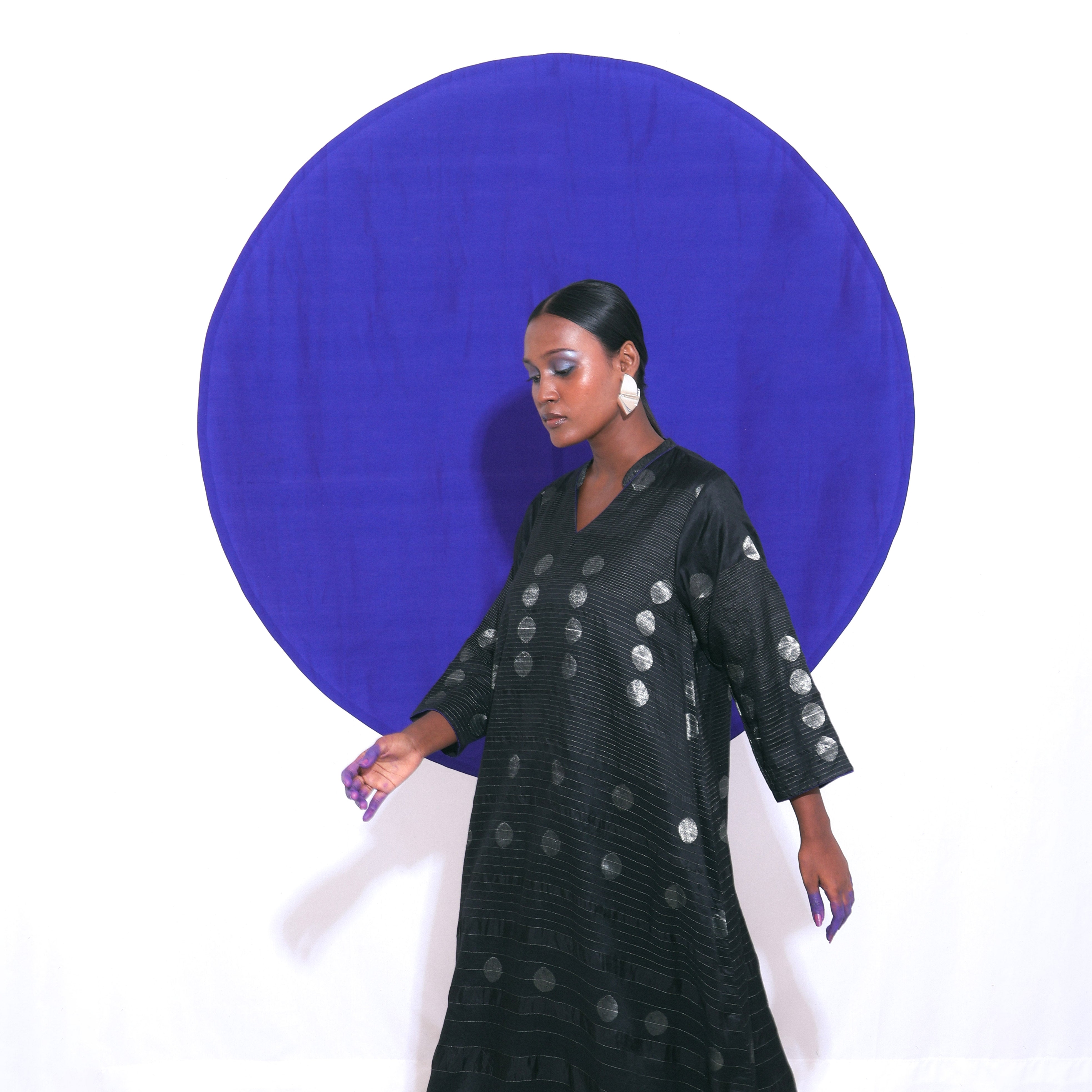 The Shoonyata Gradient Circle Flared Dress is an elegant handwoven silk piece utilizing the Jacquard Jamdani technique. It features a flattering A-line silhouette that extends to a midi length, complemented by a modest V-neckline and wide, three-quarter-length sleeves.