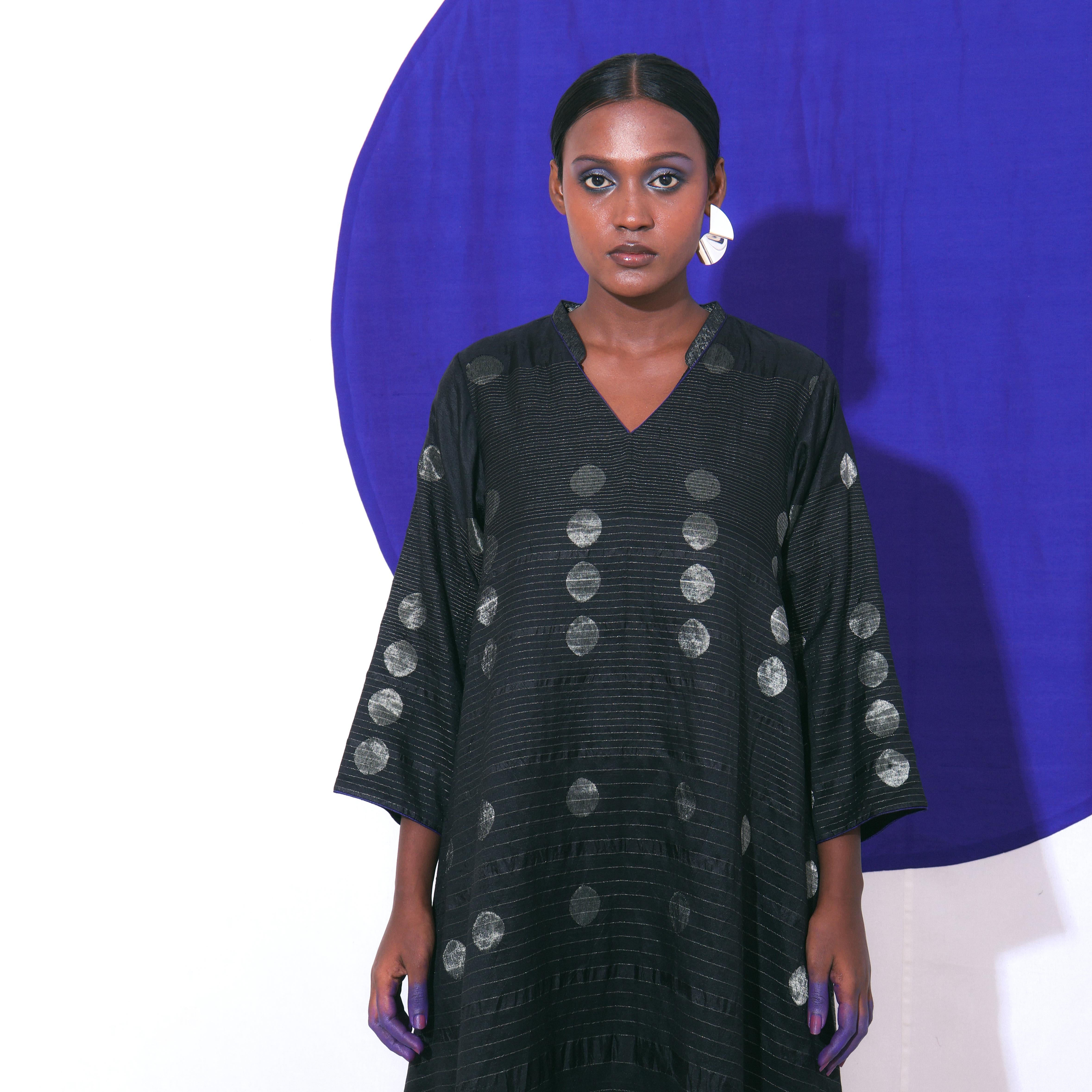 The Shoonyata Gradient Circle Flared Dress is an elegant handwoven silk piece utilizing the Jacquard Jamdani technique. It features a flattering A-line silhouette that extends to a midi length, complemented by a modest V-neckline and wide, three-quarter-length sleeves.