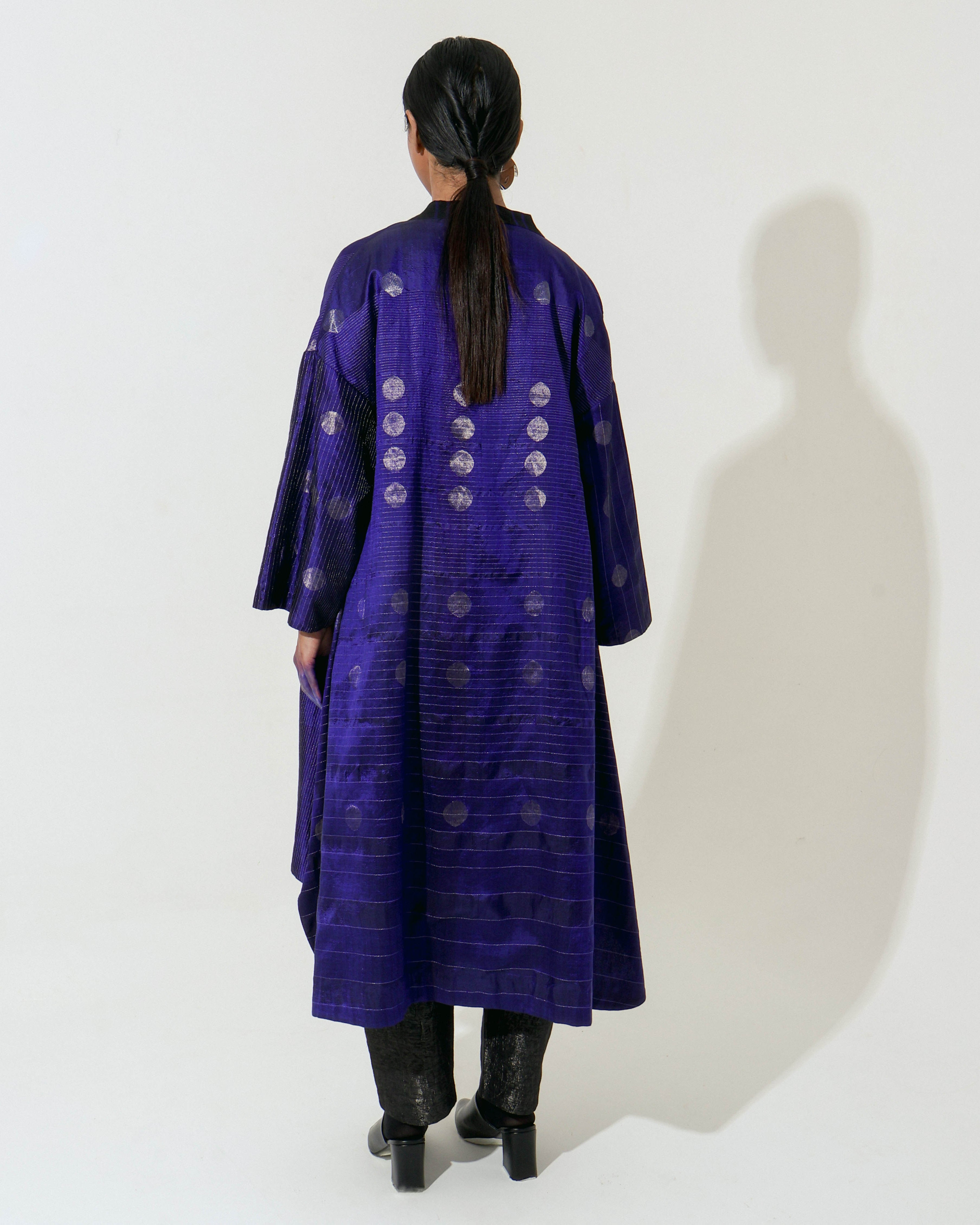 The Shoonyata Flared Kimono Jacket is a luxurious piece made from handwoven silk using the Jacquard Jamdani technique. It features intricate silver zari work and a rich royal Blue hue, enhancing its elegant appearance.
