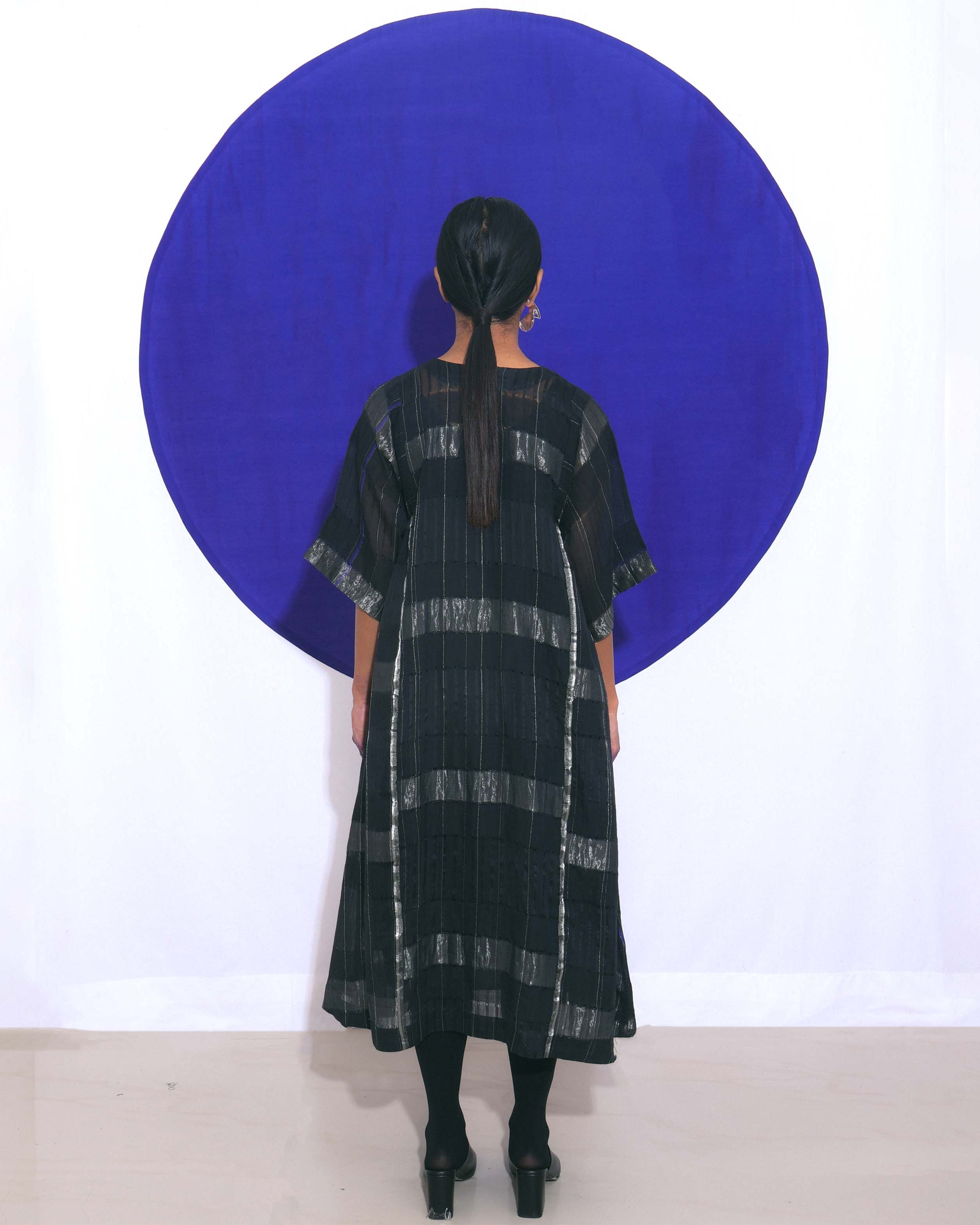 The "Shoonyata Blockstripe Panelled Dress" is an elegant dress made from handwoven silk. It features a round neckline with notched detailing, and wide, three-quarter-length sleeves for a relaxed fit.