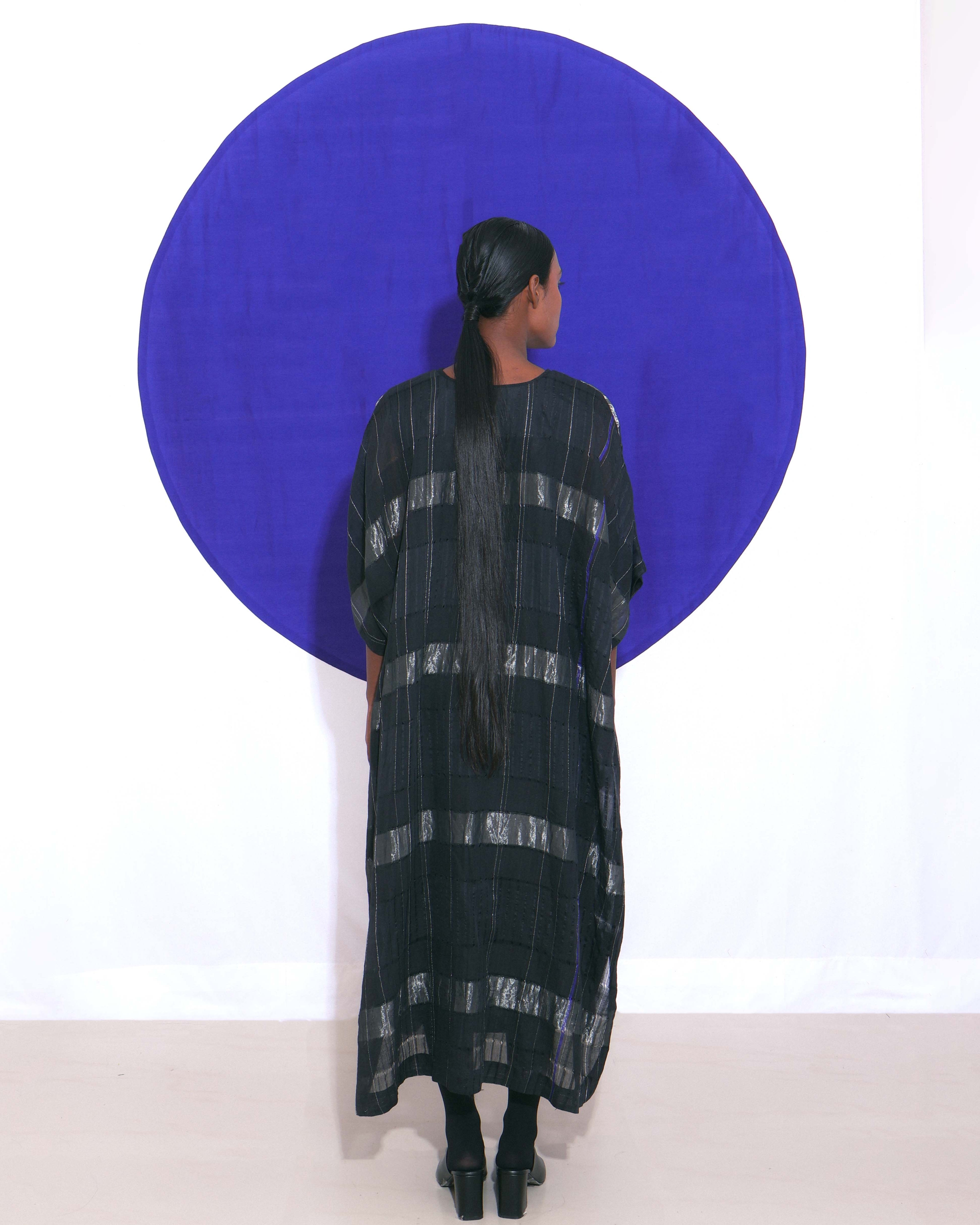 The Shoonyata Block Stripe Sheer Kaftan is a stylish, free-flowing garment made from handwoven silk. It features a relaxed, oversized silhouette, modest V-neckline, and wide elbow-length sleeves. Bold horizontal silver Zari stripes create a striking contrast against the black silk, adding a touch of luxury.