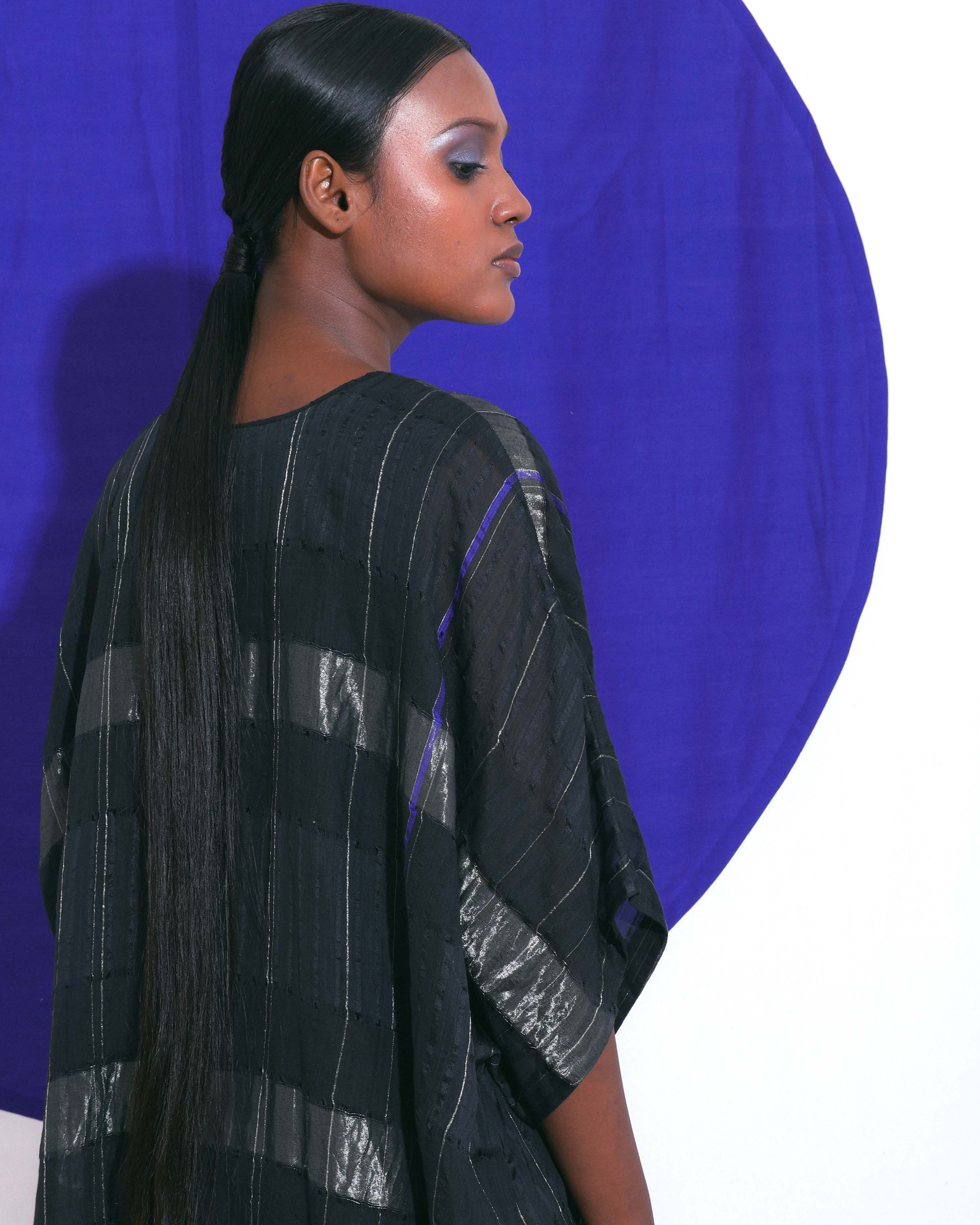The Shoonyata Block Stripe Sheer Kaftan is a stylish, free-flowing garment made from handwoven silk. It features a relaxed, oversized silhouette, modest V-neckline, and wide elbow-length sleeves. Bold horizontal silver Zari stripes create a striking contrast against the black silk, adding a touch of luxury. 
