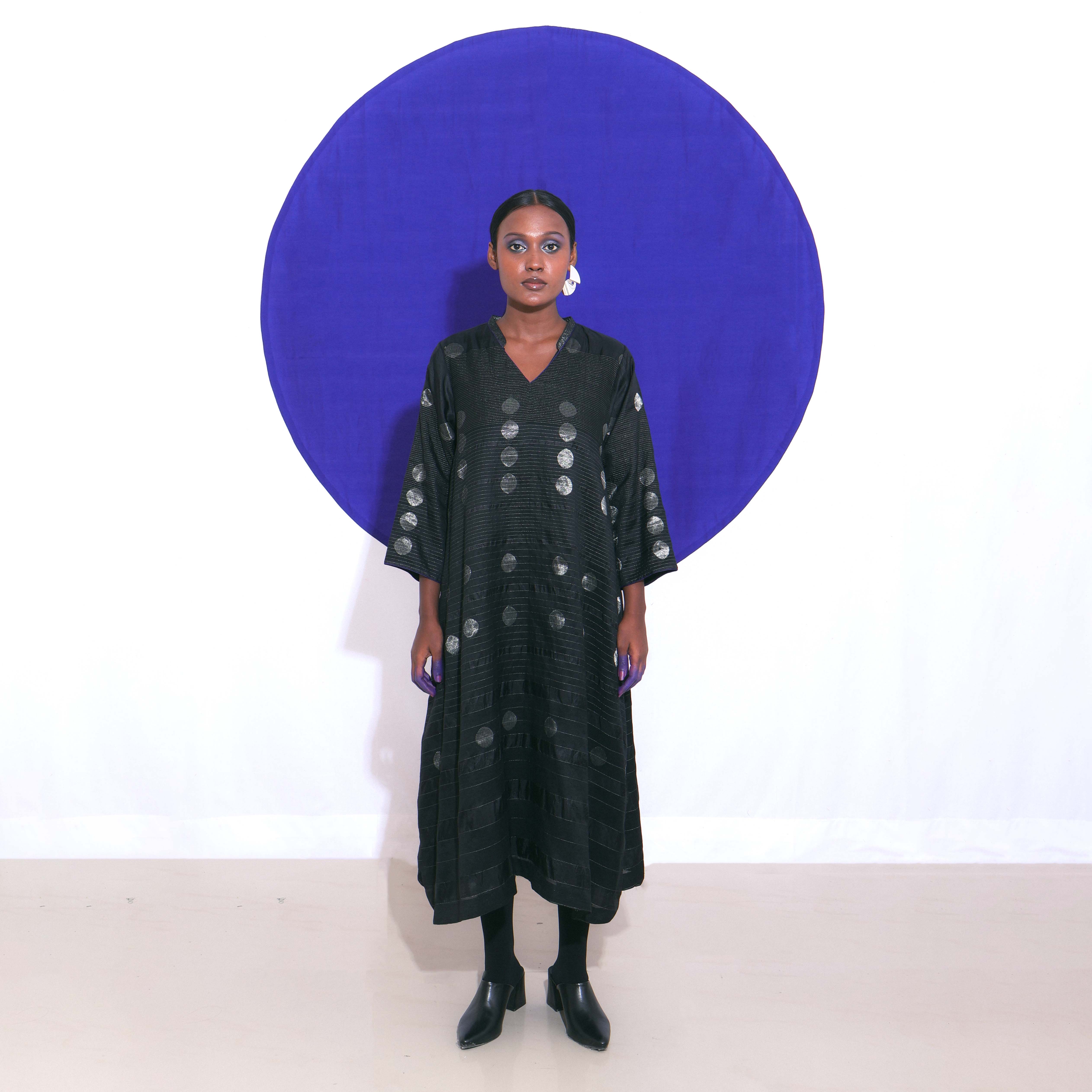 The Shoonyata Gradient Circle Flared Dress is an elegant handwoven silk piece utilizing the Jacquard Jamdani technique. It features a flattering A-line silhouette that extends to a midi length, complemented by a modest V-neckline and wide, three-quarter-length sleeves.
