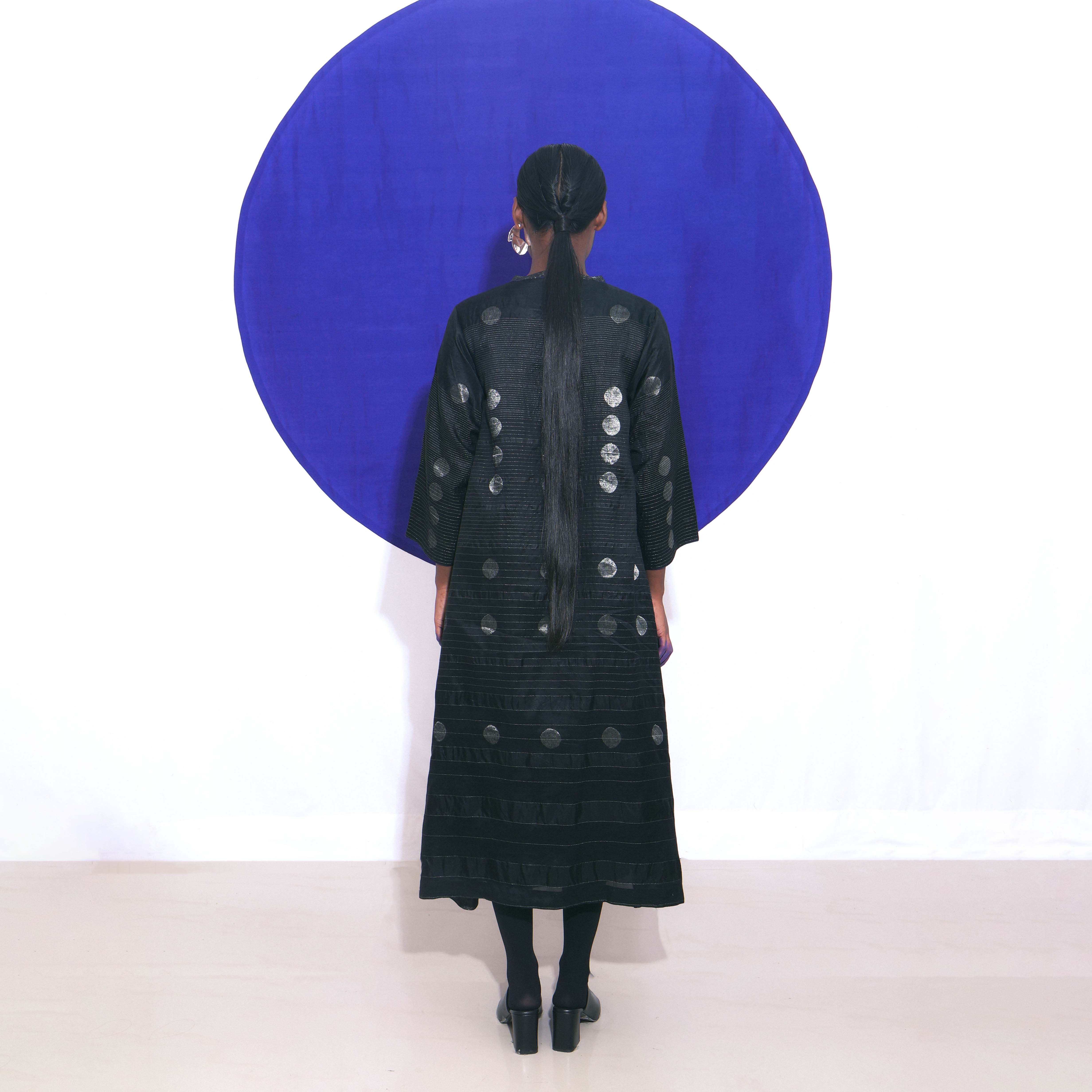 The Shoonyata Gradient Circle Flared Dress is an elegant handwoven silk piece utilizing the Jacquard Jamdani technique. It features a flattering A-line silhouette that extends to a midi length, complemented by a modest V-neckline and wide, three-quarter-length sleeves.